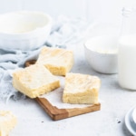 Three white chocolate brownies on a small, rectangular, wooden cutting board with a white bowl, a bottle of milk and a small bowl of white chocolate in the background.
