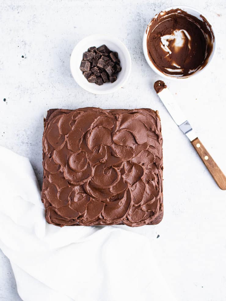 Chocolate frosted square cake on a gray backdrop with an offset spatula, a bowl of frosting and a white kitchen towel around it.