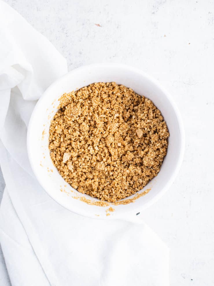 Overhead view of butter and graham cracker crumbs stirred together in a white bowl. Texture is crumbly with light clumping.