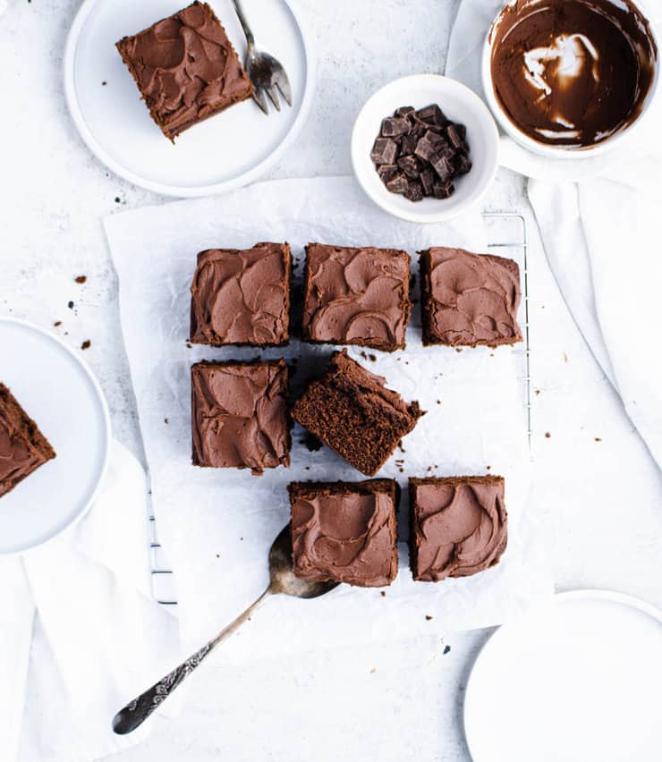 Square chocolate coffee cake cut into pieces on a parchment paper lined silver cooling rack. Plated pieces, empty plates, a bowl of chocolate and a white kitchen towel are around it.
