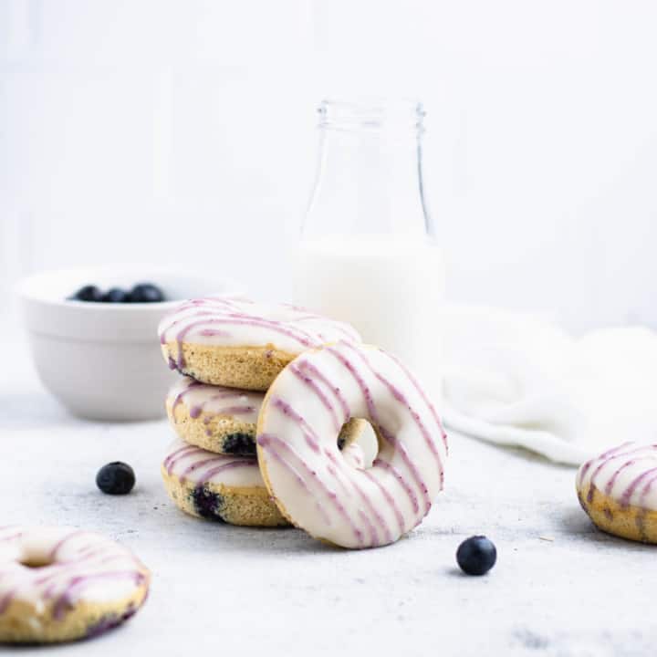 Three blueberry doughnuts stacked with another doughnut leaning against them in front of a glass of milk.