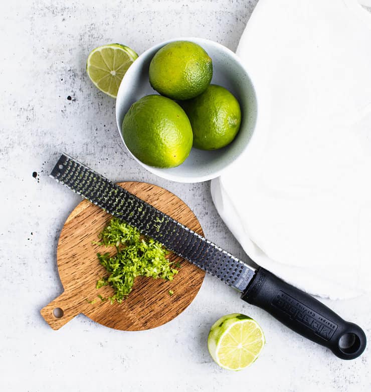 A round wooden cutting board with lime zest and a microplane on it next to a bowl of limes.