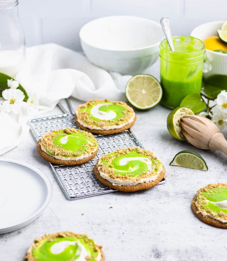 Three key lime cookies on a silver cooling rack next to a jar of lime curd, squeezed limes and an empty white plate.