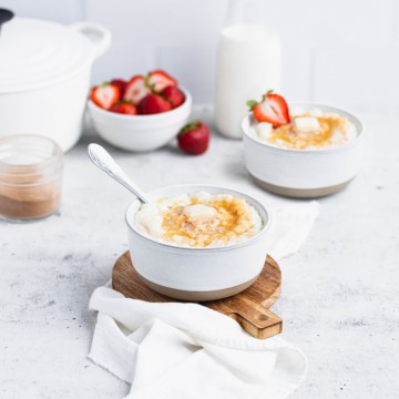 Risgrøt in a white bowl on a small round cutting board with a white napkin under it. A bowl of strawberries, another bowl of porridge, a glass of milk and a white casserole dish are in the background.