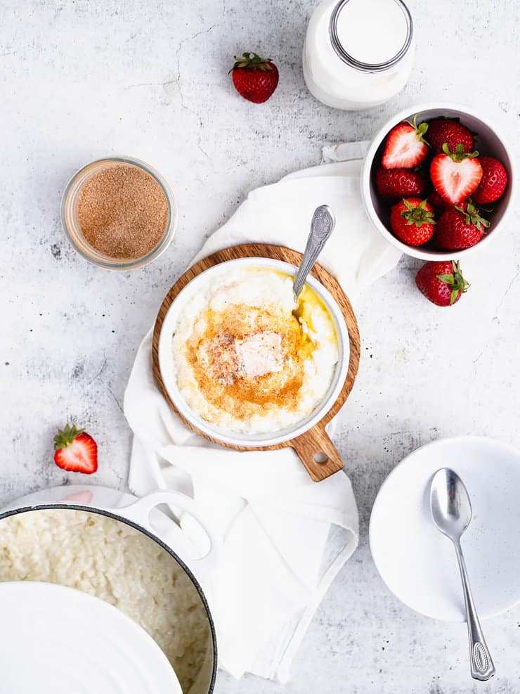 Overhead view of risgrøt in a bowl with a cube of butter and cinnamon/sugar. Around it are a bowl of strawberries, an empty bowl with a spoon and a white dutch oven with more porridge.