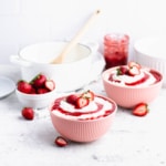Two pink bowls filled with riskrem and topped with strawberry sauce and sliced berries. In the background are a jar of sauce, a small bowl of strawberries, a white mixing bowl and a white enamel pot.
