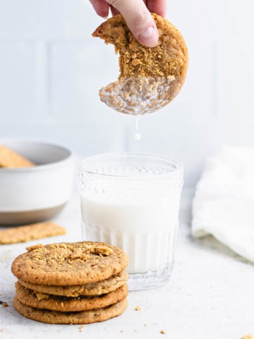 A stack of graham cracker cookies next to a glass of milk. A hand from the top of the frame is holding a cookie over the glass as milk drips off it.