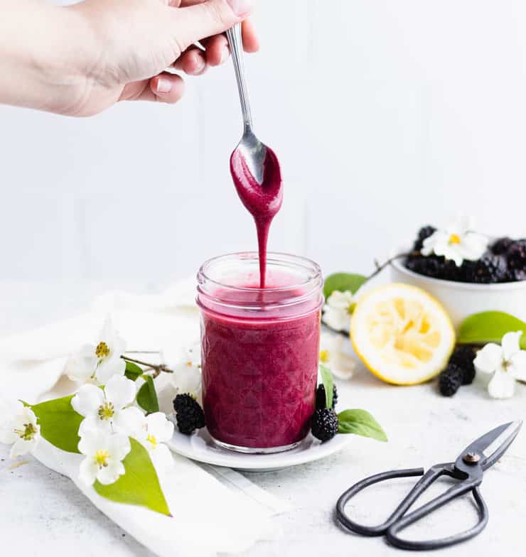 A jar of blackberry curd sits on a white plate with flowers and berries around it. A hand is holding a silver spoon over it with curd dripping off back into the jar.