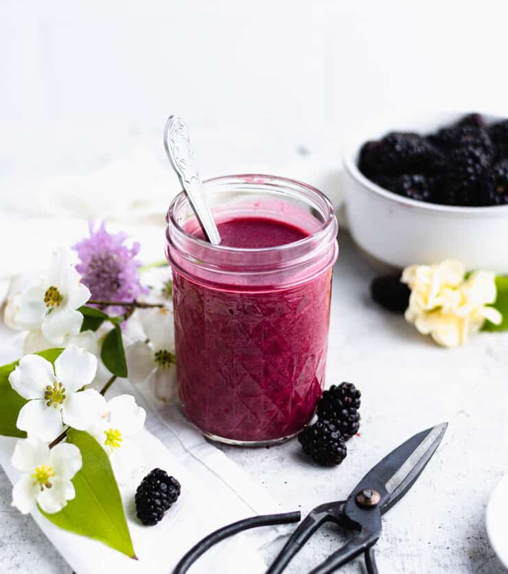 Jar of blackberry curd with a silver spoon sticking out of the top. Berries, flowers and a pair of floral scissors are laid out around it.