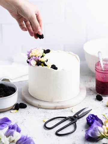 A hand placing a blackberry on top of a white frosted blackberry cake with blackberry curd, fresh blackberries, flowers and a pair of scissors around it.
