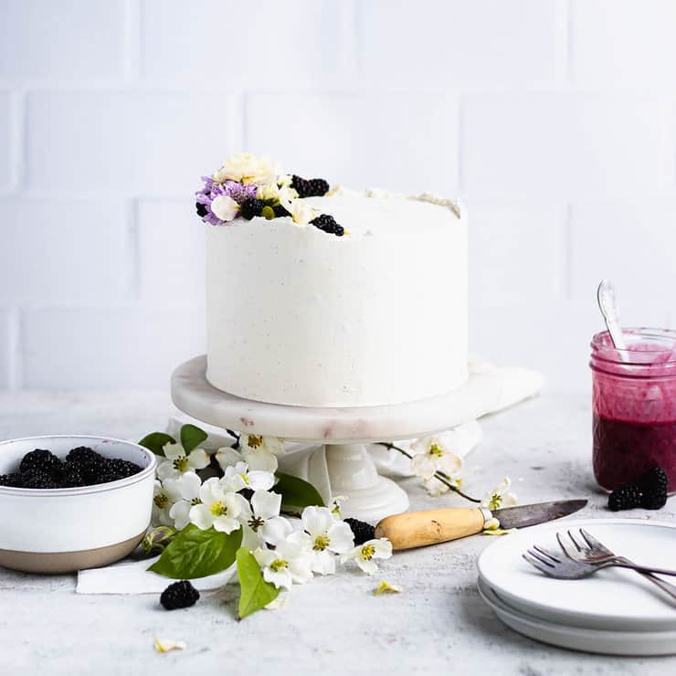 Blackberry cake on a marble cake stand with white flowers, a small knife, plates, a bowl of blackberries and a jar of blackberry curd in the background.