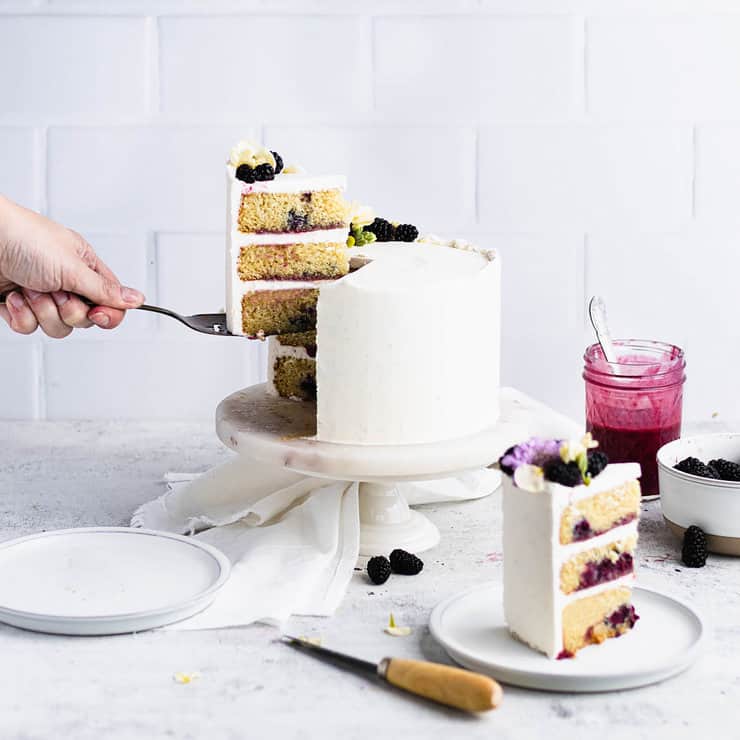 Hand pulling a slice of blackberry cake out to show the layers of vanilla cake, blackberry curd and frosting.