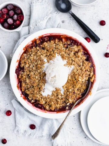 Cranberry apple crisp with a scoop of melting ice cream on a light blue striped towel. Plates, a bowl of frozen cranberries, and ice cream scoop and a white bowl are nearby.