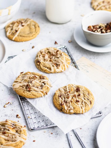 Three maple cookies on a small parchment paper-lined silver cooling rack. Around them are a glass of milk, a bowl of glaze, plates and a small bowl of pecans.