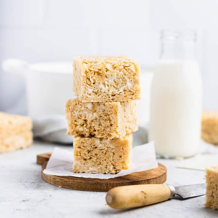 Three browned butter rice krispie treats stacked on top of each other on a small, round wood cutting board. A bottle of milk, other rice krispie treats and a white pot are in the background.