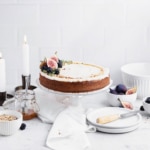 Frosted spice cake on a clear cake stand with while bowl, a jar of caramel, two taper candles and a small stack of white plates in the background.