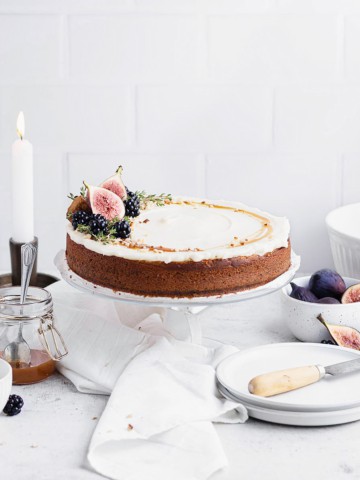 Frosted spice cake on a clear cake stand with while bowl, a jar of caramel, two taper candles and a small stack of white plates in the background.