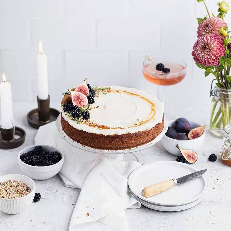 Single layer kyrdderkake decorated with a drizzle of caramel sauce, chopped nuts, blackberries, two fig halves and sprigs of thyme. In the background are bowls with these ingredients, two taper candles, a vase of deep purple dahlias and a cocktail.
