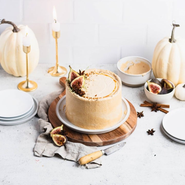 Chai cake topped with figs, anise pods, cinnamon sticks and a circle of caramel. Cake is on a white plate on a brown, round cutting board.